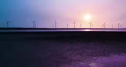 A serene landscape features numerous wind turbines on the horizon, silhouetted against a vivid sunset sky with hues of purple, pink, and blue. The turbines are reflected in a body of water below, with a dark and textured foreground adding depth to the scene.
