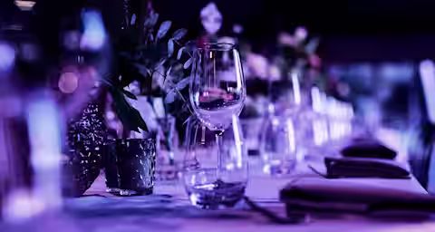 A close-up of a beautifully set dining table under purple lighting. The table features empty wine glasses, folded napkins, and small flower arrangements in vases. The background is softly blurred, creating a dreamy and elegant atmosphere.