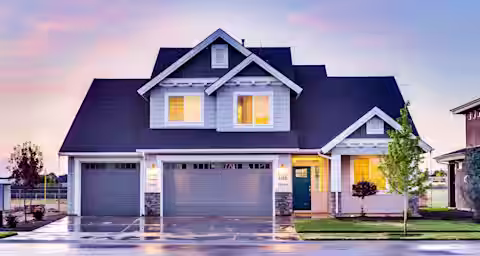 A modern two-story house with white and blue exterior, three-car garage, and a teal front door. Warm interior lights are on, and a small tree is planted in the front yard. The sky is colorful, indicating either sunrise or sunset. The driveway is slightly wet.