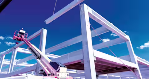 A construction site with a partially built concrete structure and a bright blue sky in the background. There is a crane lifting a concrete beam while a worker operates a cherry picker on the left side of the image.