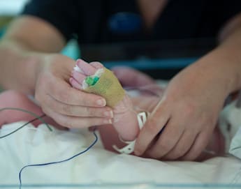 A close-up of an adult hand gently holding the foot of a newborn baby in a hospital. The baby's foot is wrapped in gauze and has medical wires attached, indicating it is receiving medical care. The adult's hand is providing a comforting touch.