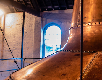 A large copper brewing kettle is seen inside a dimly lit brewery. The room has white brick walls and a large arched window in the background, illuminating the space with natural light. The ceiling is wooden with exposed beams.