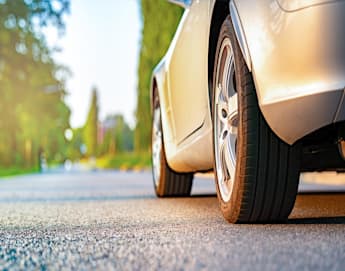 Close-up view of a car's tire on a sunlit road with blurred green trees and bushes in the background. The scene has a warm, serene atmosphere, and the road stretches out ahead.