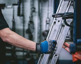 A person wearing a black shirt and a blue work glove is holding a metal ladder in a garage or workshop. Out-of-focus tools and equipment are visible in the background, indicating an industrial or mechanical setting.