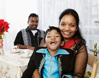 A woman and child, both smiling, sit closely together at a table with a floral tablecloth. The child wears a bright blue traditional outfit. A man, also smiling, is seated in the background. A vase with red flowers and teacups are on the table.