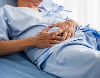 A person lying on a hospital bed wearing a light blue medical gown, with hands resting over their abdomen. The scene suggests a hospital or clinical setting with natural light filtering in from the background.