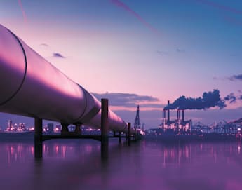 A large oil pipeline extends across a body of water towards an industrial complex with smokestacks emitting smoke. The sky is at dusk, painted in shades of purple and pink, with the water reflecting the colors and lights of the structures.