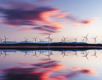 A serene landscape at sunset with wind turbines lined up on the horizon, silhouetted against a colorful sky with pink, purple, and blue hues. The scene is reflected perfectly in calm water in the foreground, creating a symmetrical and peaceful image.