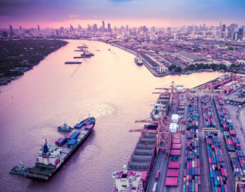 Aerial view of a large port with numerous shipping containers stacked neatly alongside large cranes. A river flows next to the port with a ship docked nearby. In the background, a sprawling city skyline is visible under a purple and pink sky.