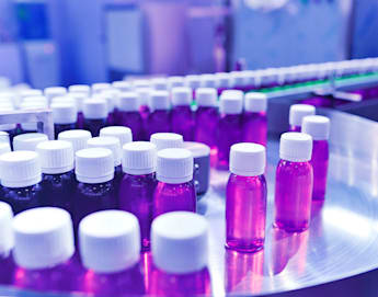 Rows of small, clear bottles filled with purple liquid are lined up on a metal circular conveyor belt in a manufacturing facility. Each bottle has a white cap. The background is a blur of industrial equipment and blue lighting, suggesting an automated process.