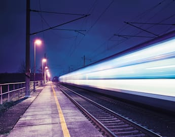 A long-exposure photograph of a train station at night, capturing the motion blur of a high-speed train passing by. The platform is illuminated by streetlights. Overhead power lines are visible against the dark blue sky.