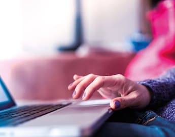 A close-up photo of a person using a laptop. The person’s right hand is typing on the keyboard. They have colorful nail polish and are wearing a long-sleeve sweater. The background is blurred, making it hard to discern specific details.