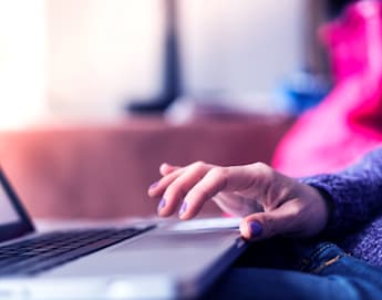 Close-up of a person's hand with purple nail polish resting on the trackpad of an open laptop. The background is softly blurred with pink and blue tones. The person appears to be using the laptop, which is situated on a flat surface.