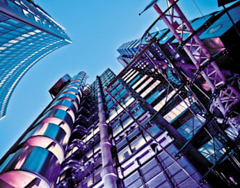 A dramatic low-angle shot of futuristic skyscrapers against a clear blue sky. The buildings, featuring sleek glass facades and metallic structures, are illuminated with vibrant purple and blue lights, emphasizing their modern architectural designs.