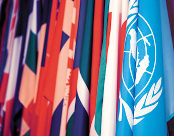 A collection of various national flags is displayed, prominently featuring the blue flag of the United Nations with its white emblem. The flags are closely arranged, showcasing a colorful and diverse array.