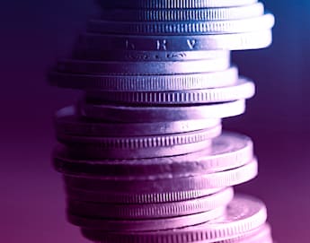 A stack of coins precariously balanced, viewed up close and illuminated with dramatic blue and purple lighting, creating a vivid contrast and giving the coins a metallic sheen. The background is blurred, keeping the focus on the coins.