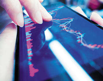 Close-up of a person's hands using a smartphone to view a graph with red and white lines and bars. The graph likely represents financial data or stock markets. The background is blurry, suggesting the person is in a busy or moving environment.