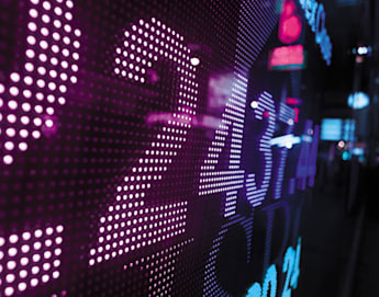 A close-up of a digital display with brightly lit numbers in various shades of purple and blue, possibly representing stock market data or financial information. The background is slightly out of focus, with blurry lights and neon-colored signs in an urban setting.