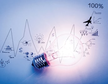 A glowing light bulb lies on a paper with various hand-drawn business-related icons, including graphs, a money bag, gears, and a plane. The background shows a blue-tinted zigzagging line graph, leading to the text "100%" at the top right corner.