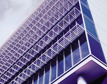 A modern, multi-story building with a sleek glass facade and grid-like metal shades covering the windows, viewed from a low angle against a clear, blue sky with a few wispy clouds.