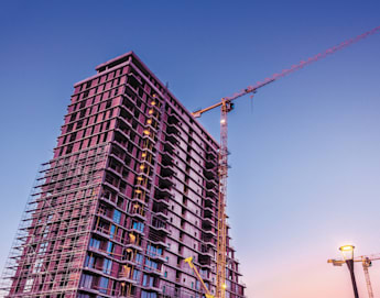 A high-rise building under construction during sunset, with cranes and scaffolding surrounding the structure. The sky is a gradient of soft purple and pink hues. A street lamp is visible in the foreground, and another crane is seen in the background.