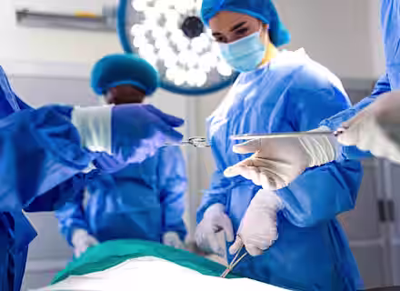 Surgeons in blue scrubs and masks perform an operation in a brightly lit operating room, with one person handing surgical scissors to another.