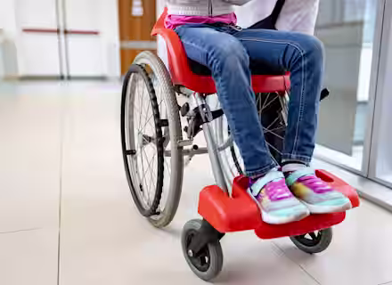 A child in a red wheelchair is being pushed by an adult down a hospital corridor. The child is wearing sneakers, jeans, and a colorful jacket, and the adult's hand is visible on the wheelchair handle.
