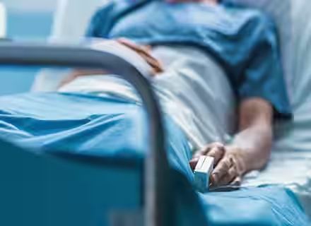 A hospital patient in a blue gown lies in a bed, partially covered by a white blanket. The patient's hand is visible, resting on the blanket, with a pulse oximeter clipped to one of their fingers.