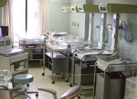 A hospital nursery room with several empty baby bassinets lined up. Each bassinet has a transparent cover and is equipped with basic monitoring devices. The room is softly lit, and curtains cover the windows partially.