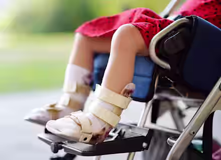 A child wearing a red dress sits in a wheelchair, with both feet in white orthopedic braces and resting on the footrests. The background is blurred and outdoors.