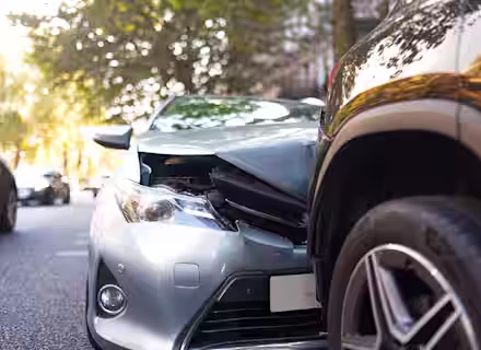 A silver car with a damaged front bumper has collided with the rear side of a black SUV on a city street lined with trees.