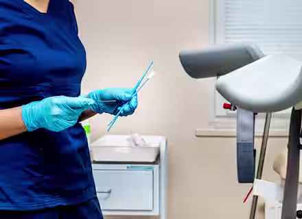 A healthcare professional in blue scrubs and gloves holds a cervical swab and brush in a medical examination room near a gynecological exam chair.