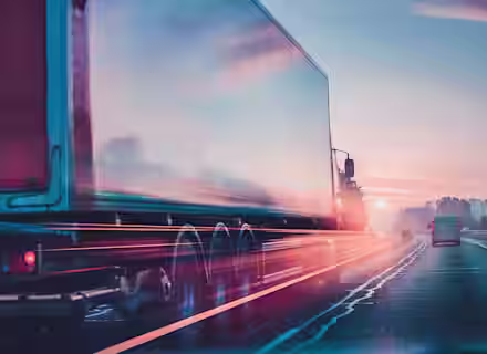 A long-exposure shot of trucks moving on a highway at sunrise or sunset. The motion blur creates streaks of light behind the vehicles, emphasizing speed. The sky is a mix of pink, orange, and blue hues, reflecting off the road and the trucks.