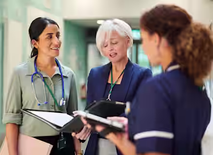 Three healthcare professionals stand in a hospital corridor, engaged in conversation. One has a stethoscope around her neck, another holds a clipboard, and the third is dressed in a nurse's uniform, holding a tablet.