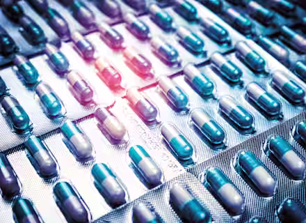 Close-up of several blister packs filled with blue and white capsules. The capsules are arranged in a grid-like pattern, and the lighting highlights some of the rows, giving a metallic sheen to the packaging.