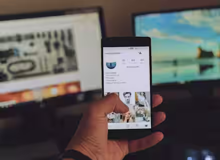 A person holds a smartphone showing an Instagram profile. In the background, there are two computer monitors; the left screen displays a technical diagram and the right screen shows a scenic beach landscape. The surroundings are dimly lit.