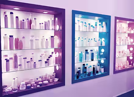 Three illuminated display shelves, each differently colored (purple, blue, and pink), showcasing various bottles, jars, and containers commonly used for cosmetics or personal care products. The items are neatly arranged, creating a visually appealing presentation.