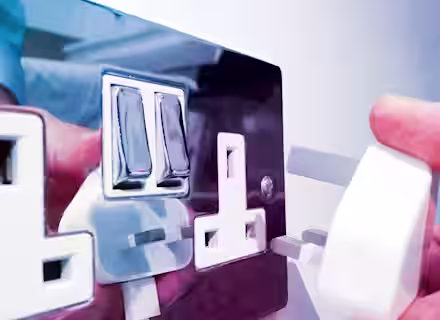 A close-up image of a person’s hand plugging a white UK three-pin plug into a shiny, metallic electrical socket on a white wall. The socket features dual switches, and their hand and the plug are reflected on the surface.