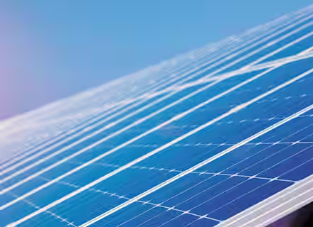 A close-up view of a large solar panel array under a clear blue sky with the sun shining brightly in the background. The solar panels reflect the sunlight, highlighting their grid-like structure and sleek surface.