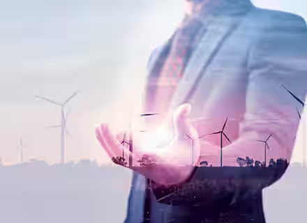 A double exposure image featuring a person in a suit holding a glowing light in their hand, superimposed with a landscape of wind turbines at sunset, symbolizing the connection between business and renewable energy.