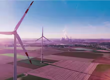 Aerial view of two large wind turbines standing in a vast, open field with a clear blue sky. Farmland stretches out in neat, colorful plots below, while industrial structures with smokestacks are visible on the horizon.