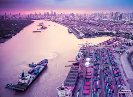 Aerial view of a large port with numerous shipping containers stacked neatly alongside large cranes. A river flows next to the port with a ship docked nearby. In the background, a sprawling city skyline is visible under a purple and pink sky.