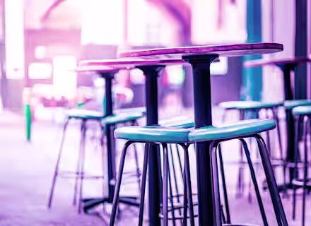 A close-up of tall bar stools and high tables in a modern indoor setting. The image is bathed in a purple and pink hue, creating a vibrant and stylish ambiance. The background is slightly blurred, emphasizing the seating area in the foreground.