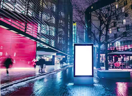 A brightly lit, modern urban plaza at night with buildings on both sides. In the center, there is a large blank digital billboard. People are walking around, and a few trees and outdoor seating areas are visible. The ground is wet, reflecting the lights.