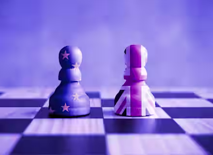 Two chess pawns face each other on a chessboard. The left pawn features the European Union flag design, while the right pawn is adorned with the United Kingdom flag. The image is tinted with a blue hue, symbolizing the Brexit theme.