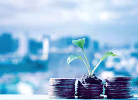 A small plant grows out of a pile of soil on top of a stack of coins, set against a blurred cityscape background. The scene symbolizes financial growth and investment in sustainability.