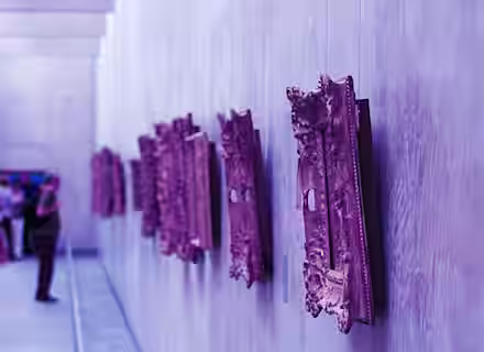 An art gallery with ornate, purple-hued frames mounted on the wall. The frames extend out towards the viewer, showing their intricate detailing. The gallery's corridor stretches into the distance, with a few people blurred in the background.