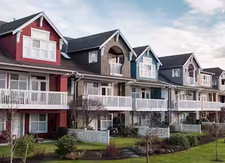 A row of stylish, multi-colored houses with large windows, spacious balconies, and gabled roofs, all lined up side by side. Each house has a small front yard with a white picket fence, and bare trees and cloudy sky can be seen in the background.