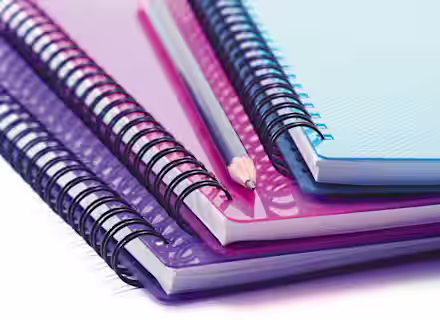 A stack of colorful spiral-bound notebooks lies on a flat surface, featuring blue, purple, and pink covers. A sharpened pencil rests diagonally across the top notebook. The notebooks are slightly askew, creating a layered, overlapping effect.