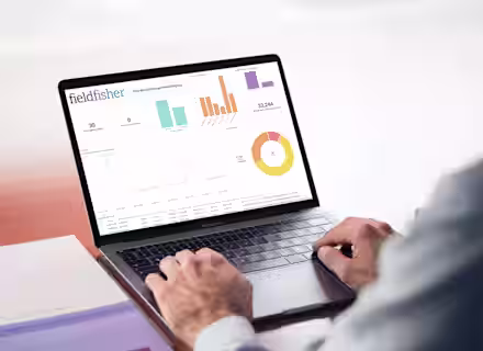 A person types on a laptop displaying various colorful graphs and charts under the heading "fieldfisher." The screen shows bar graphs, a line graph, and a pie chart, emphasizing data analysis. The person's face is not visible.